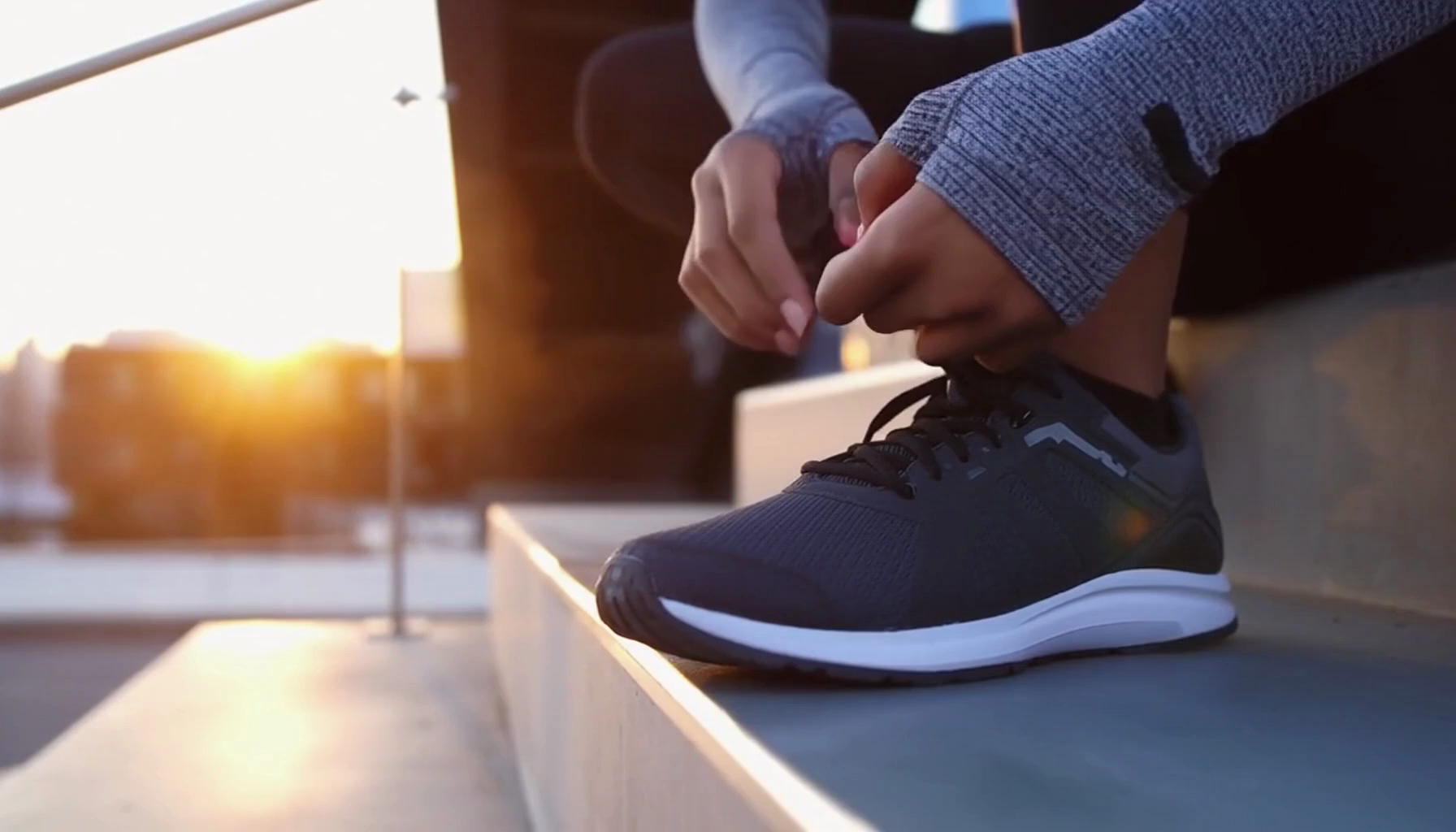 A slow‑motion lifestyle commercial shot of a runner tying their shoes on a modern concrete staircase at dawn. Crisp reflections, realistic fabric physics, subtle breath vapor in the cold air, drifting dust particles, and clean cinematic lighting.